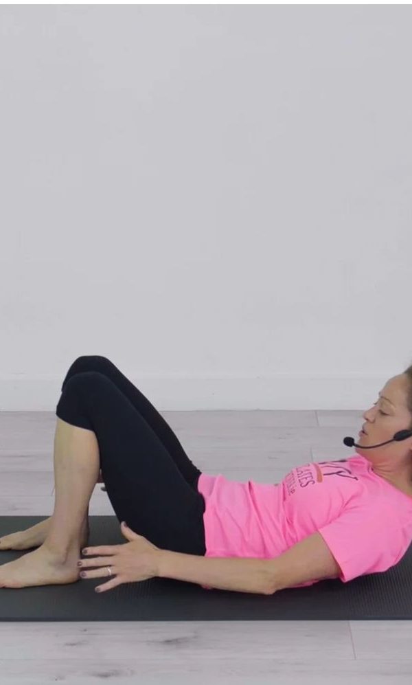 Woman, pink t-shirt, grey mat, demonstrating a Pilates exercise
